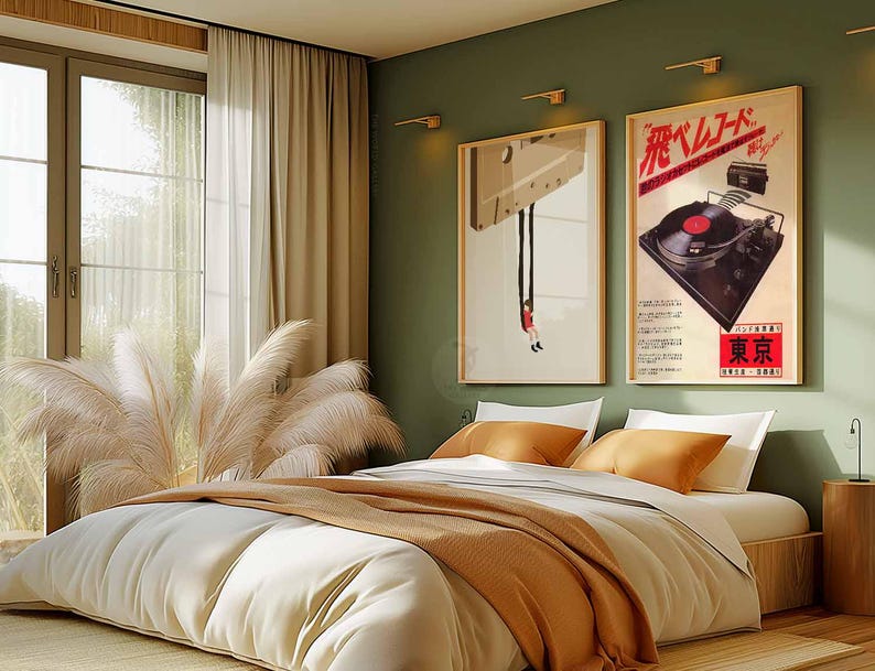 Op de afbeelding: Een slaapkamerinterieur met een bed, een raam en twee ingelijste posters aan de muur. Beide posters zijn vintage advertenties voor platenspelers. De poster aan de linkerkant toont een platenspeler die aan het plafond hangt met een persoon die eronder op de grond zit. De poster aan de rechterkant toont een platenspeler met de tekst "飛 レ コ ー ド" in het Japans. Het bed is opgemaakt met witte lakens en een bruine deken. Er liggen twee kussens op het bed, een wit en een bruin.