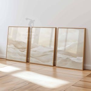 Könnte beinhalten: Drei gerahmte abstrakte Kunstdrucke in einem beige-weißen Farbschema. Die Drucke zeigen ein wellenförmiges, fließendes Design, das an Wolken oder Sanddünen erinnert.