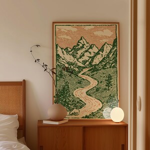 May include: A framed landscape art print featuring a mountain range, river, and green valley. The artwork is displayed above a wooden cabinet with a round vase, books, and a spherical lamp. The color palette includes green, beige, and brown tones.