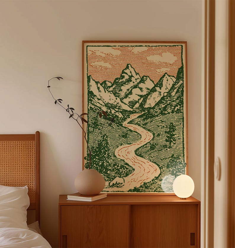 May include: A framed landscape art print featuring a mountain range, river, and green valley. The artwork is displayed above a wooden cabinet with a round vase, books, and a spherical lamp. The color palette includes green, beige, and brown tones.