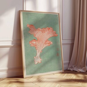 May include: A framed art print featuring a coral-colored mushroom illustration on a muted teal background. The artwork is displayed in a light wooden frame, leaning against a white wall and wooden floor, with a beige curtain in the background.