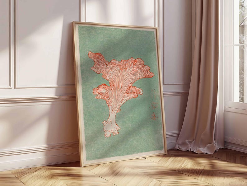 May include: A framed art print featuring a coral-colored mushroom illustration on a muted teal background. The artwork is displayed in a light wooden frame, leaning against a white wall and wooden floor, with a beige curtain in the background.