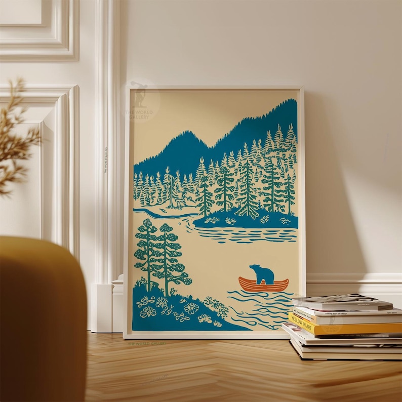May include: A framed art print featuring a stylized illustration of a forest scene. The artwork depicts a bear in an orange canoe on a lake, surrounded by teal trees and mountains against a cream background. The print is displayed on a wooden floor.