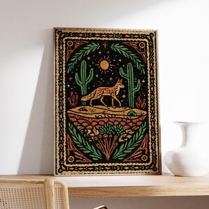May include: A framed art print featuring a desert scene with a fox, cacti, and sun. The artwork has a black background with orange, green, and yellow accents. The print is displayed on a wooden shelf.