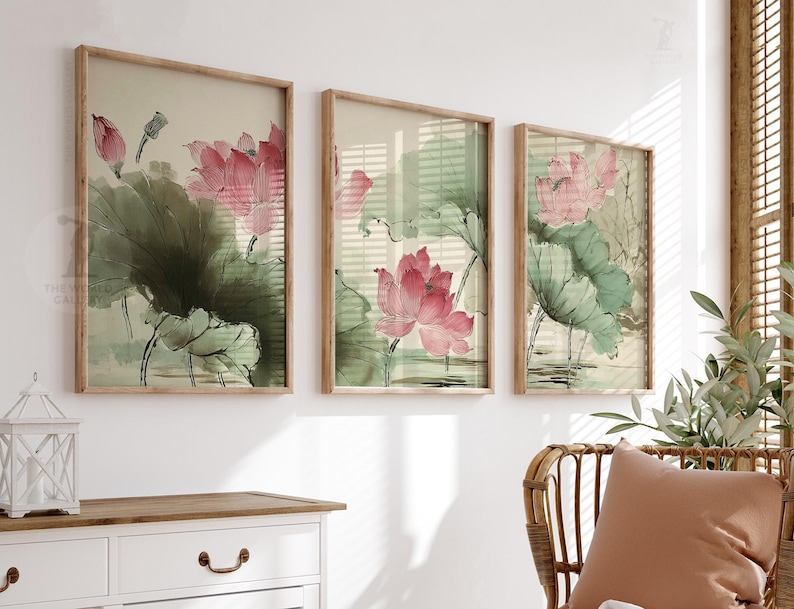 May include: Three framed art prints featuring watercolor paintings of pink lotus flowers and green leaves. The artwork has a vintage aesthetic, with a light beige background and wooden frames. The prints are displayed above a white dresser.