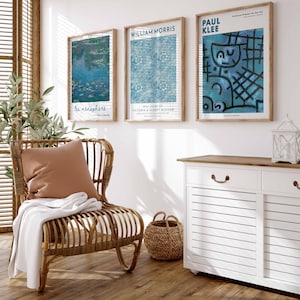May include: Three framed art prints on a white wall, featuring works by Monet, William Morris, and Paul Klee. A wicker chair with a brown cushion and white throw is in front of a white cabinet with a wooden top. A woven basket sits on the wooden floor.