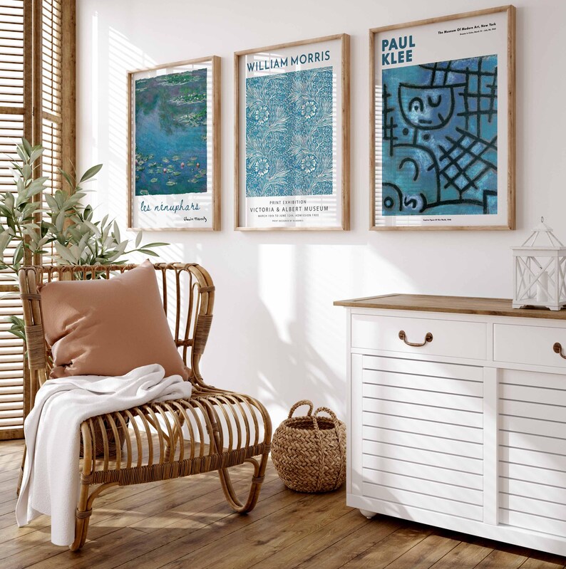 May include: Three framed art prints on a white wall, featuring works by Monet, William Morris, and Paul Klee. A wicker chair with a brown cushion and white throw is in front of a white cabinet with a wooden top. A woven basket sits on the wooden floor.