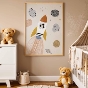 May include: A framed art print featuring a whimsical rocket ship with a cat in the cockpit, surrounded by planets and stars. The artwork is in a child's bedroom, alongside a white dresser, teddy bears, and a crib.