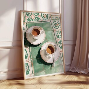 May include: A framed art print depicts two white teacups with coffee on saucers, resting on a green and white tiled surface. The artwork is leaning against a white wall and a wooden floor, with a beige curtain and window in the background.