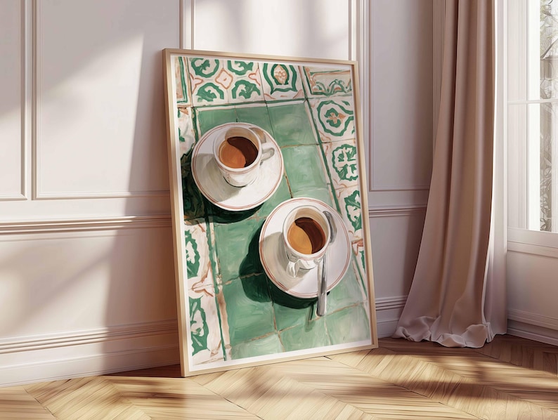 May include: A framed art print depicts two white teacups with coffee on saucers, resting on a green and white tiled surface. The artwork is leaning against a white wall and a wooden floor, with a beige curtain and window in the background.