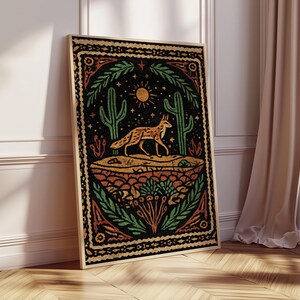 May include: A framed art print featuring a desert scene with a fox, cacti, and sun. The artwork has a black background with orange, green, and yellow accents. The print is displayed on a wooden floor, leaning against a white wall.