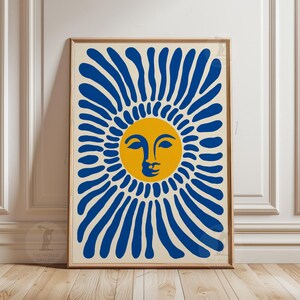 May include: A framed art print featuring a stylized sun with a yellow face and blue rays against a cream background. The artwork is in a wooden frame and is displayed against a white wall.