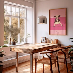 May include: A wooden desk with metal legs and two woven chairs. A framed print of a cow on a pink sofa hangs on the wall. A large window with a view of trees is in the background.