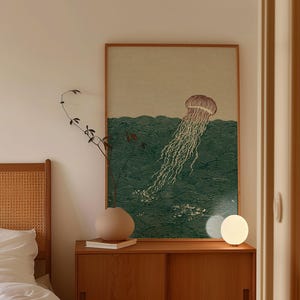 May include: A framed print featuring a jellyfish in a sea of green waves. The artwork has a vintage aesthetic, with a light brown frame. A small, round, white lamp sits on a wooden cabinet below the print.