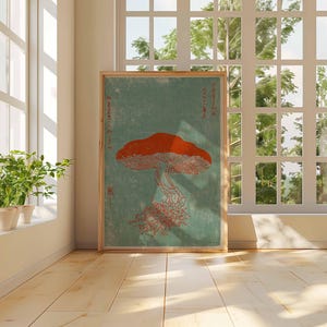 May include: A framed art print featuring a stylized orange mushroom with a detailed cap and stem against a teal background. The artwork is displayed in a bright room with a window and potted plants, creating a natural aesthetic.