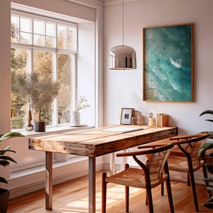 May include: A bright, airy home office with a large wooden desk and two woven chairs. A framed ocean-themed artwork hangs on the wall. A large window offers a view of the outdoors, and a modern pendant light hangs above the desk.