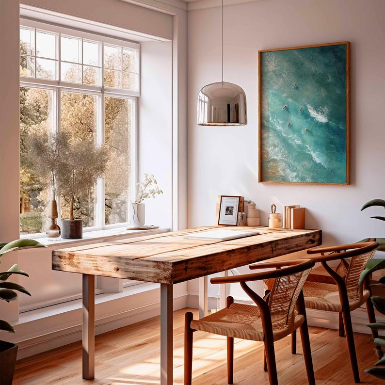 May include: A bright, airy home office with a large wooden desk and two woven chairs. A framed ocean-themed artwork hangs on the wall. A large window offers a view of the outdoors, and a modern pendant light hangs above the desk.
