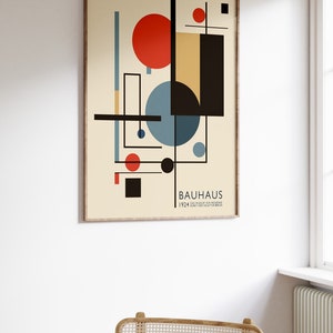 May include: A framed Bauhaus art print with geometric shapes in red, black, blue, and beige. The print includes the word "BAUHAUS" and the year "1924". The artwork is displayed on a white wall above a woven chair.