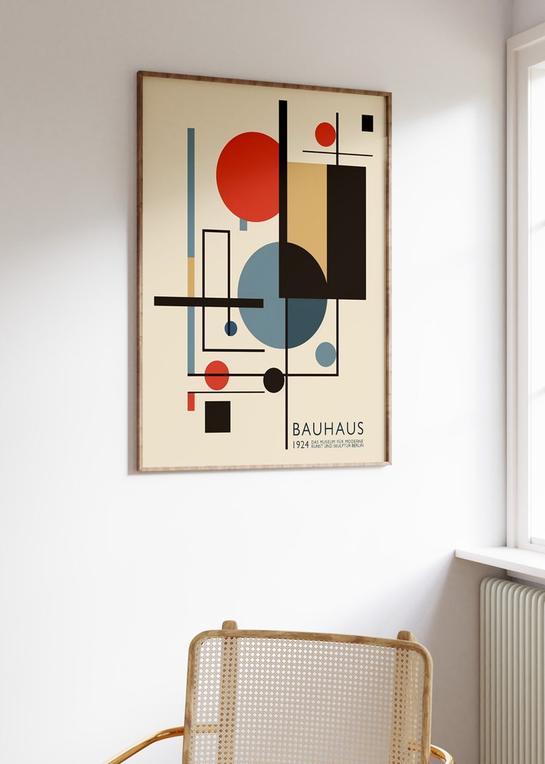 May include: A framed Bauhaus art print with geometric shapes in red, black, blue, and beige. The print includes the word "BAUHAUS" and the year "1924". The artwork is displayed on a white wall above a woven chair.