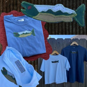 May include: Various light blue and navy blue t-shirts with a fish applique. One shirt is folded, and two are hanging on wooden hangers. A large fish applique is also displayed. The shirts are casual wear.