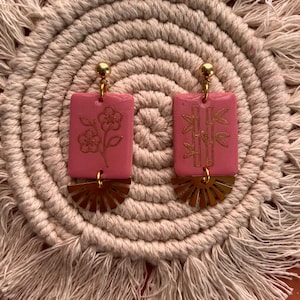 May include: Pink rectangular earrings with gold accents. One earring features a flower design, the other bamboo. Each has a gold sunburst-shaped base and a gold ball at the top. The earrings are displayed on a beige, woven surface.