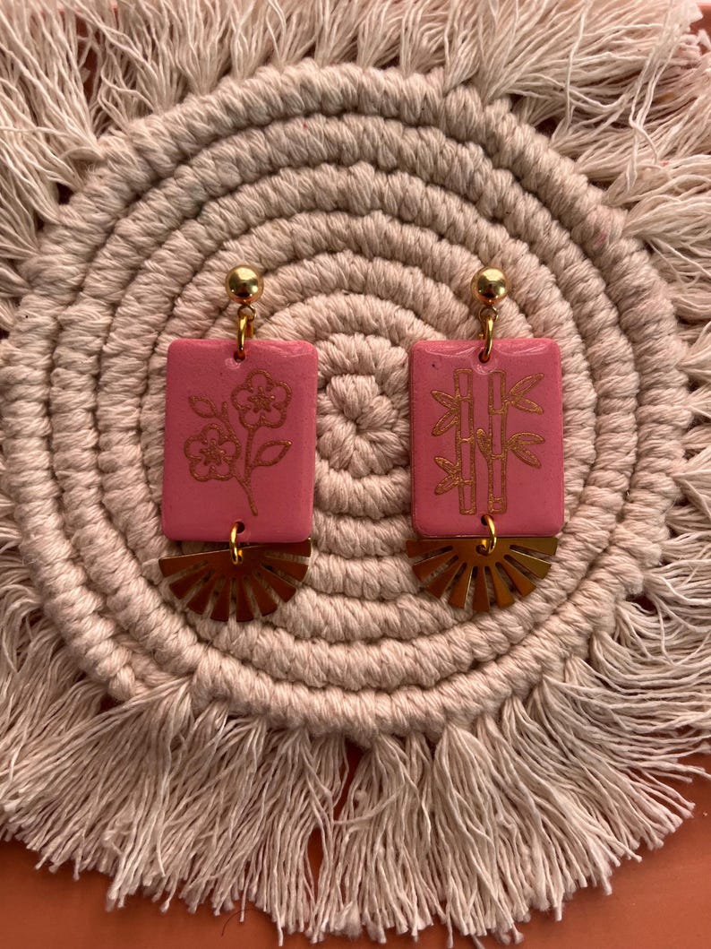 May include: Pink rectangular earrings with gold accents. One earring features a flower design, the other bamboo. Each has a gold sunburst-shaped base and a gold ball at the top. The earrings are displayed on a beige, woven surface.