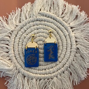 May include: A pair of blue and gold earrings. Each earring has a rectangular blue base with a gold top. One earring says "JOKER" and features a dragon design. The other earring has a dragon design.