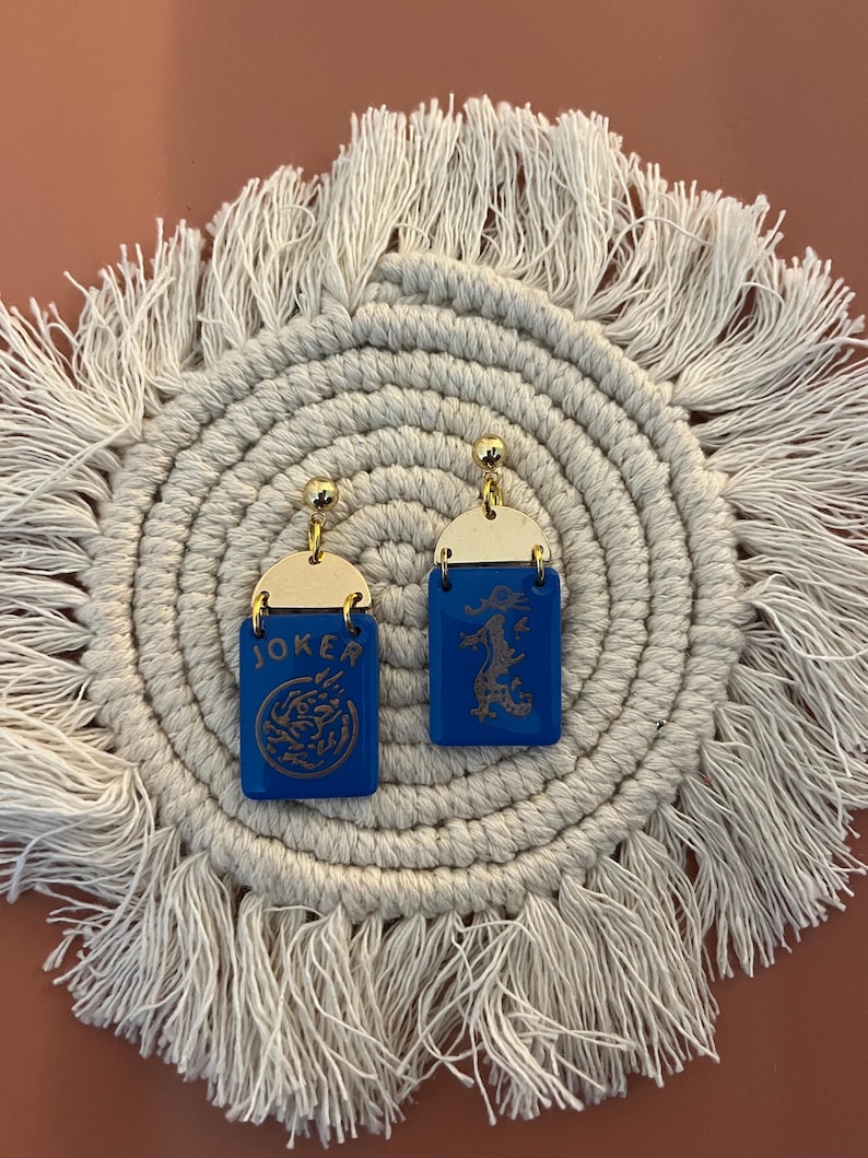May include: A pair of blue and gold earrings. Each earring has a rectangular blue base with a gold top. One earring says "JOKER" and features a dragon design. The other earring has a dragon design.