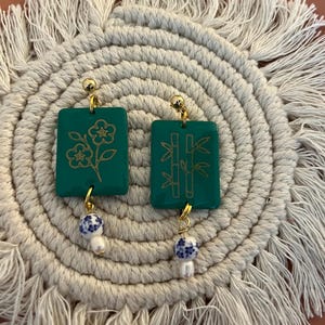 May include: A pair of green rectangular earrings with gold floral and bamboo designs. Each earring has a gold ball stud and a dangling charm with a blue and white pattern and a white pearl. The earrings rest on a cream-colored, circular woven mat.