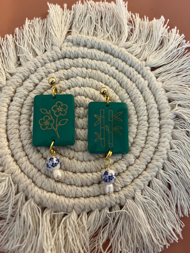 May include: A pair of green rectangular earrings with gold floral and bamboo designs. Each earring has a gold ball stud and a dangling charm with a blue and white pattern and a white pearl. The earrings rest on a cream-colored, circular woven mat.