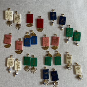 May include: A collection of rectangular earrings in various colors, including red, blue, green, and white. Each earring features a gold-colored fan-shaped accent and a small, dangling bead. Some earrings have symbols or characters.