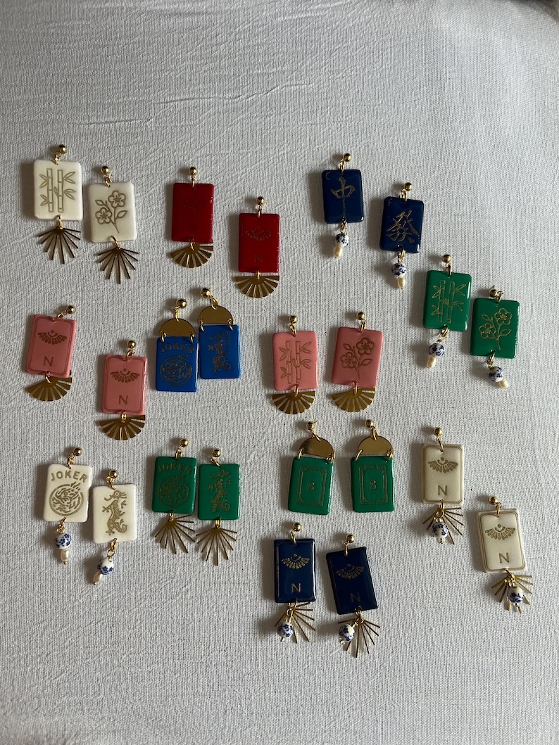 May include: A collection of rectangular earrings in various colors, including red, blue, green, and white. Each earring features a gold-colored fan-shaped accent and a small, dangling bead. Some earrings have symbols or characters.