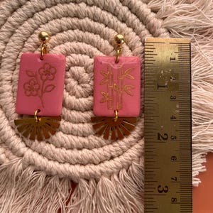 May include: Pink rectangular earrings with gold floral and bamboo designs, and gold fan-shaped accents. The earrings are approximately 1.5 inches long, with a gold ball at the top. The earrings are displayed on a beige woven mat.