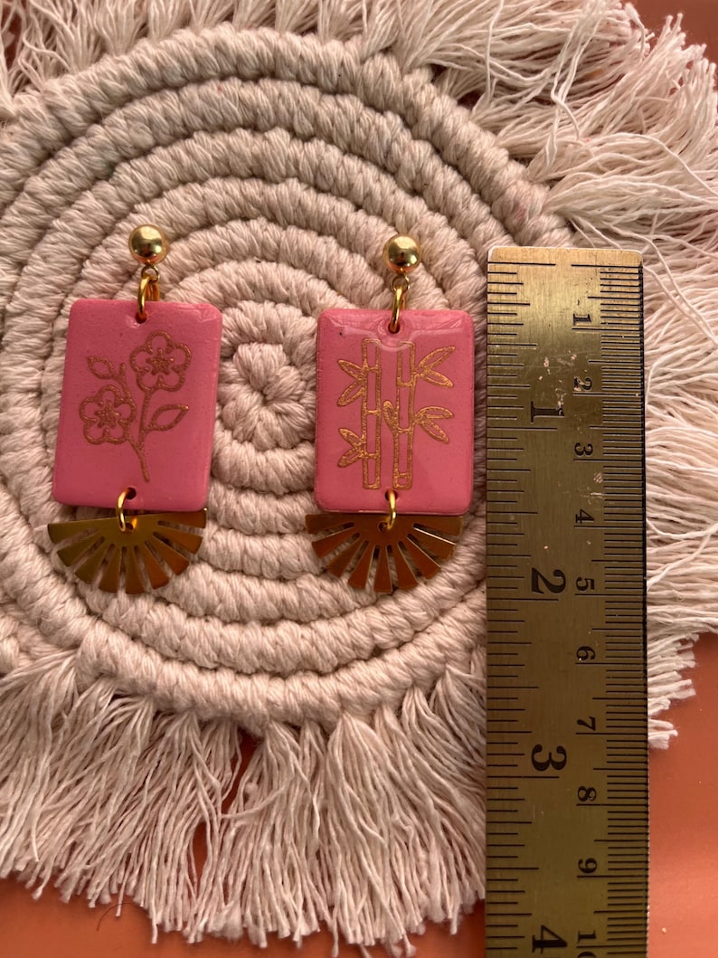May include: Pink rectangular earrings with gold floral and bamboo designs, and gold fan-shaped accents. The earrings are approximately 1.5 inches long, with a gold ball at the top. The earrings are displayed on a beige woven mat.
