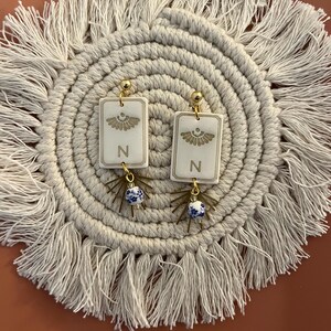 May include: A pair of gold-toned earrings with rectangular white pendants. Each pendant features a gold design and the letter "N". Below the pendant hangs a blue and white ceramic bead. The earrings are displayed on a cream-colored, fringed placemat.