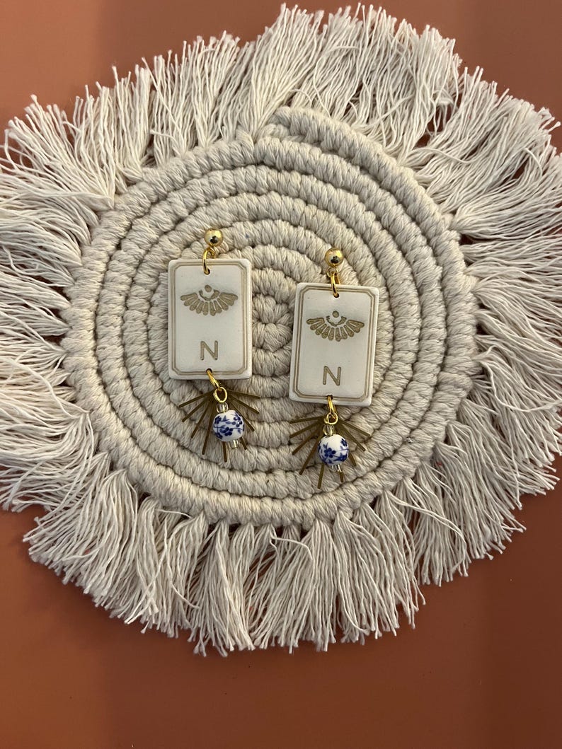 May include: A pair of gold-toned earrings with rectangular white pendants. Each pendant features a gold design and the letter "N". Below the pendant hangs a blue and white ceramic bead. The earrings are displayed on a cream-colored, fringed placemat.