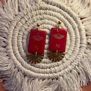 May include: A pair of red rectangular earrings with gold accents. Each earring features a gold design and the letter "N". Below the rectangle is a gold fan-shaped element. The earrings are displayed on a cream-colored, fringed placemat.
