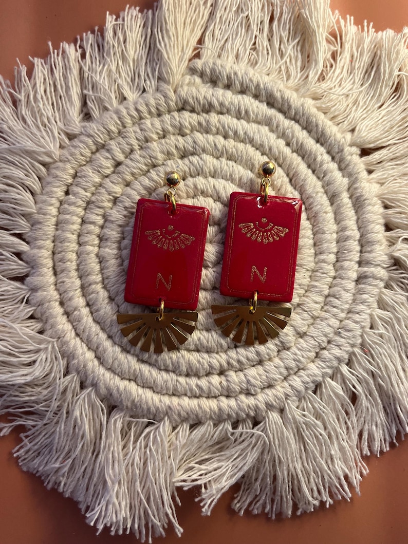 May include: A pair of red rectangular earrings with gold accents. Each earring features a gold design and the letter "N". Below the rectangle is a gold fan-shaped element. The earrings are displayed on a cream-colored, fringed placemat.