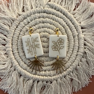 May include: A pair of white rectangular earrings with gold-colored bamboo and flower designs. The earrings have gold-colored ball studs and fan-shaped charms. The earrings are displayed on a beige, fringed placemat.