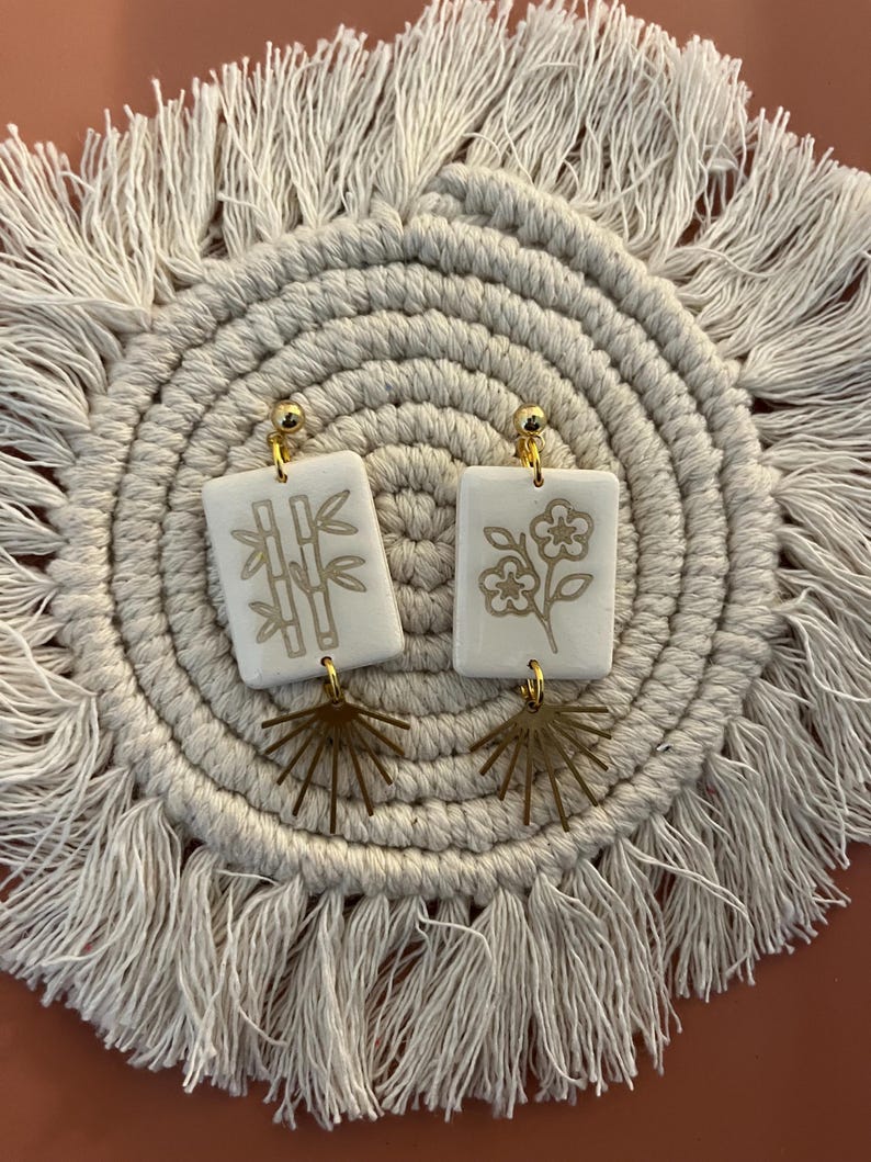 May include: A pair of white rectangular earrings with gold-colored bamboo and flower designs. The earrings have gold-colored ball studs and fan-shaped charms. The earrings are displayed on a beige, fringed placemat.