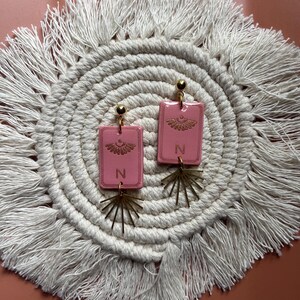 May include: A pair of pink rectangular earrings with gold-colored accents. Each earring features a gold-colored sunburst design and the letter "N". The earrings are displayed on a white macrame coaster with a fringed edge.