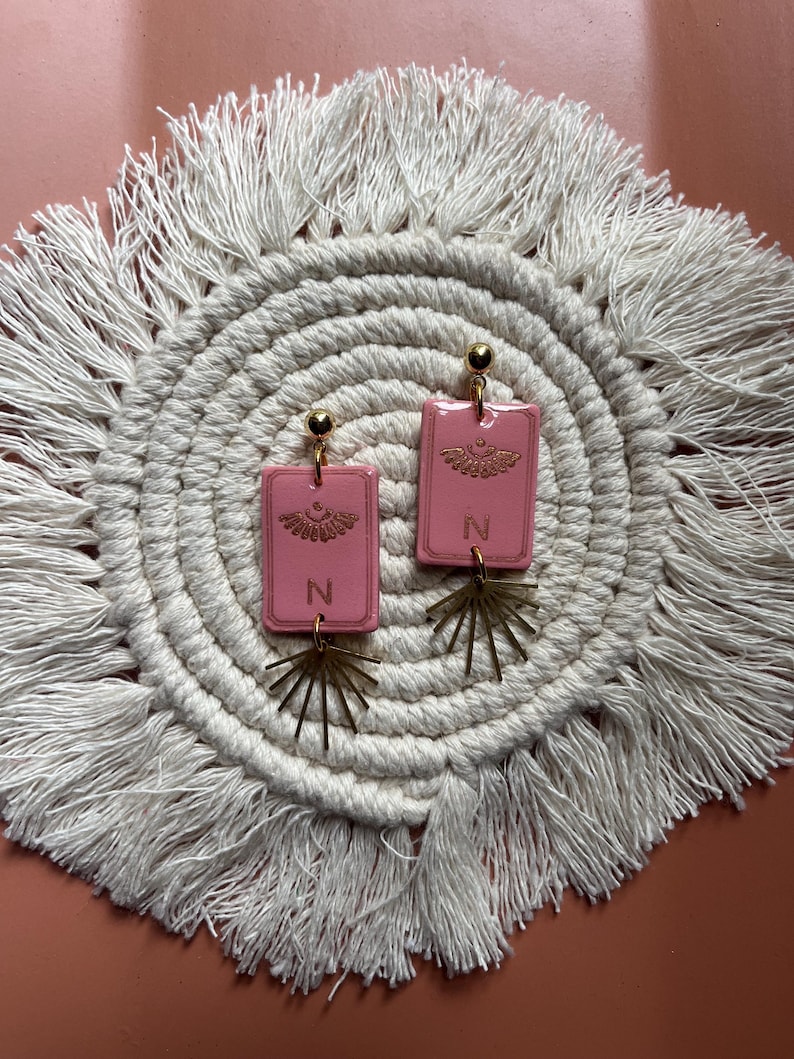 May include: A pair of pink rectangular earrings with gold-colored accents. Each earring features a gold-colored sunburst design and the letter "N". The earrings are displayed on a white macrame coaster with a fringed edge.