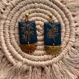 May include: A pair of teal rectangular earrings with gold bamboo and floral designs. Each earring has a gold sunburst-shaped charm and a gold ball at the top. The earrings are displayed on a beige woven mat.