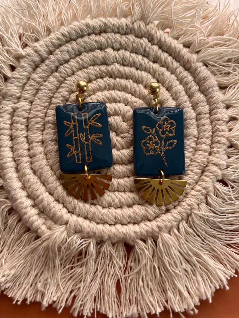 May include: A pair of teal rectangular earrings with gold bamboo and floral designs. Each earring has a gold sunburst-shaped charm and a gold ball at the top. The earrings are displayed on a beige woven mat.