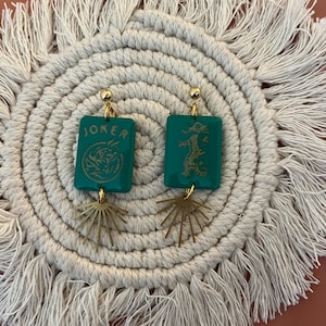 May include: A pair of teal earrings with gold accents. Each earring features a rectangular teal charm with gold text and a gold sunburst charm. One earring says "JOKER" and the other has a dragon design.