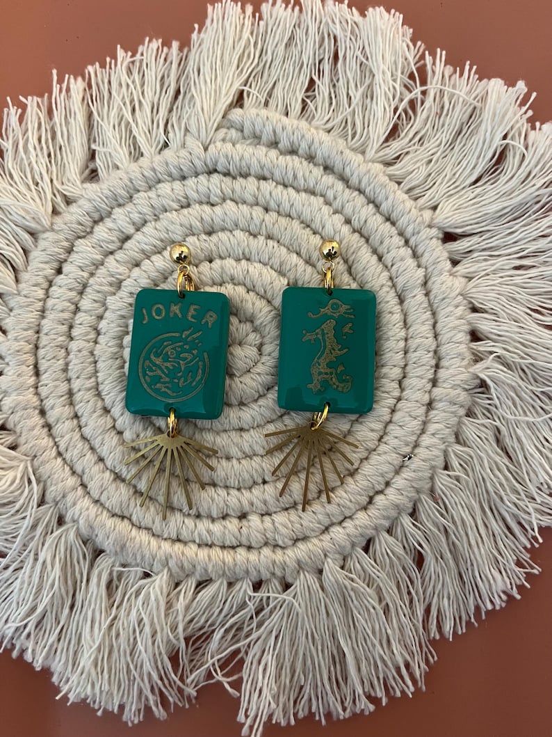 May include: A pair of teal earrings with gold accents. Each earring features a rectangular teal charm with gold text and a gold sunburst charm. One earring says "JOKER" and the other has a dragon design.