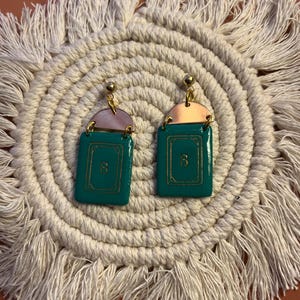 May include: A pair of teal and gold earrings with a rectangular shape, featuring the letter "B" in gold. The earrings have a gold top and a pink accent. They are displayed on a cream-colored, fringed placemat.