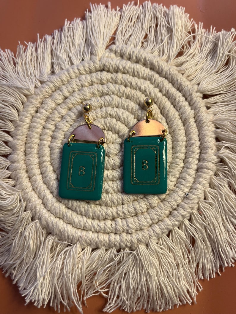 May include: A pair of teal and gold earrings with a rectangular shape, featuring the letter "B" in gold. The earrings have a gold top and a pink accent. They are displayed on a cream-colored, fringed placemat.