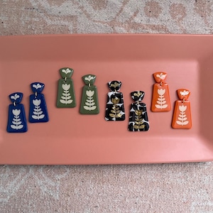 May include: A collection of handmade earrings in various colors, including blue, green, black, and orange. Each earring features a floral design in white. The earrings are displayed on a rectangular pink tray.