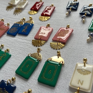 May include: A collection of colorful earrings with gold-colored accents. The earrings feature rectangular and square shapes in various colors, including red, pink, green, and blue. Some earrings have gold-colored fan-shaped details and decorative patterns.